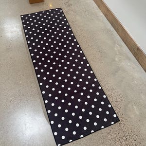 May include: A rectangular yoga mat featuring a black base color with a repeating white polka dot pattern. The mat is laid out flat, showcasing a consistent design across its entire surface, providing a clear view of the monochromatic aesthetic.