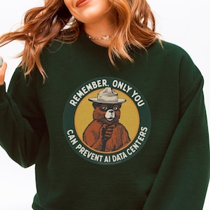 May include: A dark green long-sleeve sweatshirt featuring a circular graphic of a brown bear wearing a ranger hat and pointing forward. The text surrounding the bear reads, "REMEMBER, ONLY YOU CAN PREVENT AI DATA CENTERS" in white lettering.