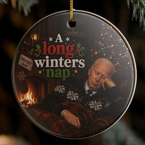 May include: Round Christmas ornament with a black background, featuring an image of a person sleeping in front of a fireplace. The ornament has the text "A long winter's nap" and is hanging from a gold string.