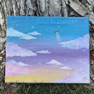 May include: Acrylic painting on canvas depicting a dreamy sky scene. The artwork features a gradient of colors, including blue, purple, pink, and yellow, with fluffy clouds, stars, and a UFO. The painting is signed in the bottom right corner.