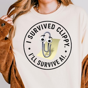 May include: Cream-colored sweatshirt with a circular graphic featuring a cartoon paperclip with googly eyes and the text "I Survived Clippy. I'll Survive AI.". The sweatshirt has long sleeves and a relaxed fit.