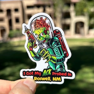 May include: A white sticker with a green alien holding a ray gun. The alien has a large head with a red brain. The sticker says "I Got My Ack Probed in Roswell, NM".