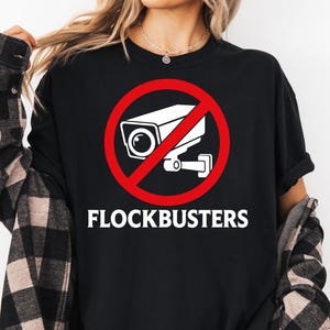 May include: A black t-shirt featuring a white graphic of a security camera inside a red prohibition circle, with the text FLOCKBUSTERS printed in white underneath. The shirt is worn with a black and white plaid long-sleeve button-down shirt.
