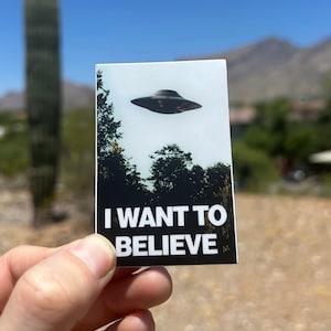 May include: A white sticker with a black and white image of a UFO and the text "I WANT TO BELIEVE".