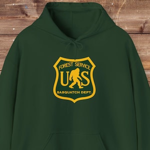 May include: A dark green hooded sweatshirt with a yellow graphic of a stylised Bigfoot inside a shield with the text "Forest Service U.S. Sasquatch Dept."