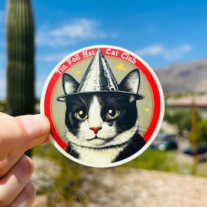 May include: A round sticker with a black and white cat wearing a silver tin foil hat. The sticker has a red border and the text "Tin Foil Hat Cat Club" in white.