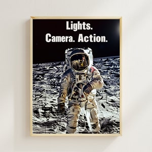 May include: A black and white photo of an astronaut standing on the moon. The astronaut is wearing a white spacesuit with a helmet and a US flag patch on the shoulder. The text "Lights. Camera. Action." is printed above the astronaut.
