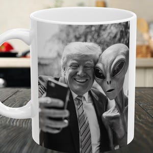 May include: White ceramic mug featuring a black and white image of a man and an alien taking a selfie. The man is smiling and holding a phone. The alien has large, dark eyes. The mug has a white handle.