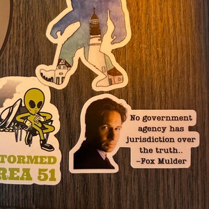 Puede incluir: Una colecci&oacute;n de pegatinas con varios dise&ntilde;os. Una pegatina presenta una silueta de Bigfoot con un faro y edificios. Otra muestra un extraterrestre con el texto "Stormed Area 51". Una tercera pegatina tiene una cita de Fox Mulder.
