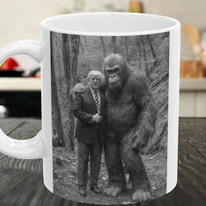 May include: White ceramic mug featuring a black and white image of a man in a suit standing with a large, hairy creature in a forest setting. The mug has a curved handle and a glossy finish, suitable for hot beverages.