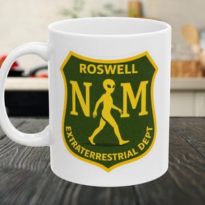 May include: A white ceramic mug featuring a green and yellow shield-shaped emblem. The graphic displays the text ROSWELL N M and EXTRATERRESTRIAL DEPT with a yellow silhouette of an alien figure walking in the center of the shield design.