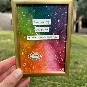 Peut inclure: Une peinture à l'aquarelle encadrée dans un cadre doré. L'œuvre présente un design de galaxie colorée avec le texte "Soar as high and as far as your dreams take you." Un petit autocollant d'OVNI est également inclus.