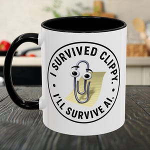 以下が含まれることがあります： 黒いハンドルと縁が付いた白いコーヒーマグ。マグカップには、クリッピーの漫画画像、ペーパークリップ、テキスト「I SURVIVED CLIPPY. I'LL SURVIVE AI.」が黒い円形デザインで表示されています。
