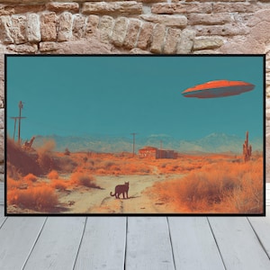 May include: A framed art print depicting a desert landscape under a teal sky. An orange UFO hovers above a dirt road, with a black cat in the foreground. The scene includes cacti, mountains, and a small building, all in warm tones.