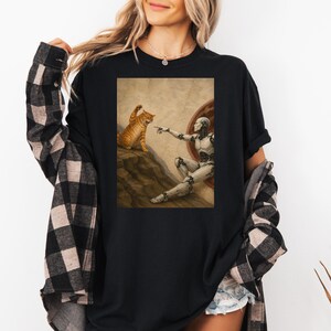 May include: Black t-shirt featuring a parody of Michelangelo's The Creation of Adam. The artwork depicts a robot and an orange tabby cat. The robot's finger is outstretched towards the cat, which is sitting on a rocky surface.