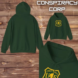May include: A dark green hooded sweatshirt with a yellow graphic of a forest service logo featuring a Bigfoot-like creature. The logo reads "Forest Service US" and "Investigation Dept". The text "Conspiracy Corp" is printed above the image.
