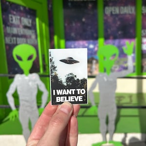Pu&ograve; includere: Una carta bianca con testo nero che dice "I WANT TO BELIEVE" con un'immagine in bianco e nero di un disco volante sullo sfondo. La carta &egrave; tenuta da una mano davanti a un muro verde con un cartello che dice "WELCOME TO ROSWELL OPEN DAILY".