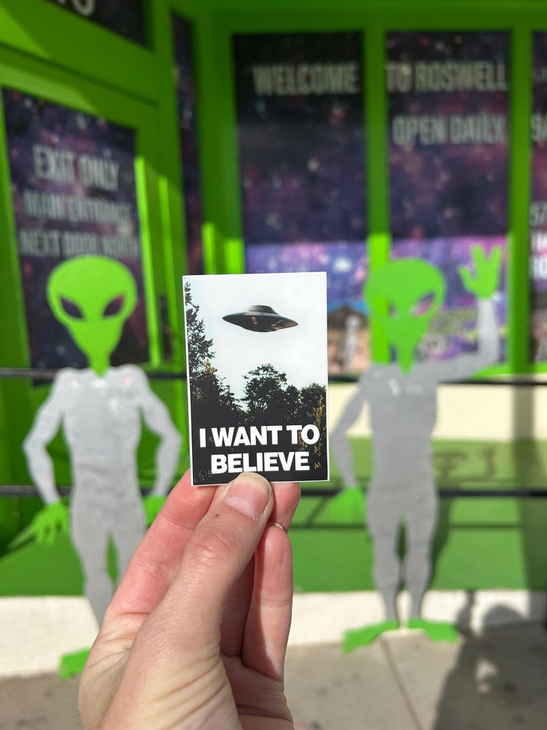 Pu&ograve; includere: Una carta bianca con testo nero che dice "I WANT TO BELIEVE" con un'immagine in bianco e nero di un disco volante sullo sfondo. La carta &egrave; tenuta da una mano davanti a un muro verde con un cartello che dice "WELCOME TO ROSWELL OPEN DAILY".