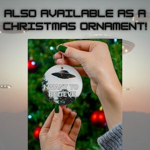 Pu&ograve; includere: Un ornamento natalizio in ceramica bianca con un'immagine in bianco e nero di un UFO e il testo "I WANT TO BELIEVE". L'ornamento &egrave; appeso a un filo dorato ed &egrave; tenuto da una mano.