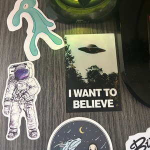 Pu&ograve; includere: Una collezione di adesivi che presentano una figura aliena blu, un astronauta con una galassia nel suo casco, un UFO in bianco e nero con il testo "I WANT TO BELIEVE", un'illustrazione in bianco e nero di un drago e una figura mascherata, e un adesivo in bianco e nero con il testo "Biz"