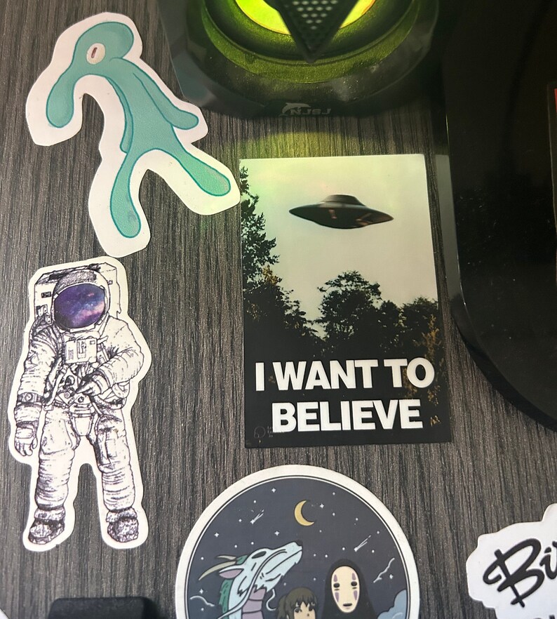 Pu&ograve; includere: Una collezione di adesivi che presentano una figura aliena blu, un astronauta con una galassia nel suo casco, un UFO in bianco e nero con il testo "I WANT TO BELIEVE", un'illustrazione in bianco e nero di un drago e una figura mascherata, e un adesivo in bianco e nero con il testo "Biz"
