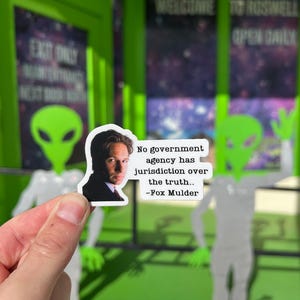 Puede incluir: Una pegatina con un retrato de Fox Mulder y la cita: "Ninguna agencia gubernamental tiene jurisdicci&oacute;n sobre la verdad". La pegatina se sostiene frente a un recorte de un extraterrestre verde y un edificio con el letrero "Welcome to Roswell".