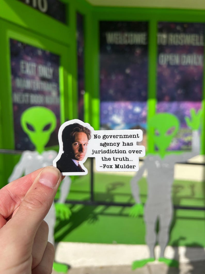 Puede incluir: Una pegatina con un retrato de Fox Mulder y la cita: "Ninguna agencia gubernamental tiene jurisdicci&oacute;n sobre la verdad". La pegatina se sostiene frente a un recorte de un extraterrestre verde y un edificio con el letrero "Welcome to Roswell".