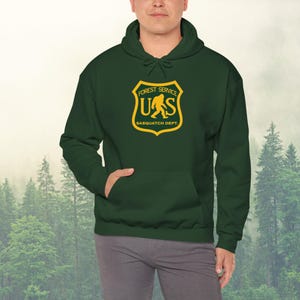 May include: A dark green hooded sweatshirt with a yellow and brown patch featuring a stylized image of a sasquatch and the text "Forest Service US Sasquatch Dept."