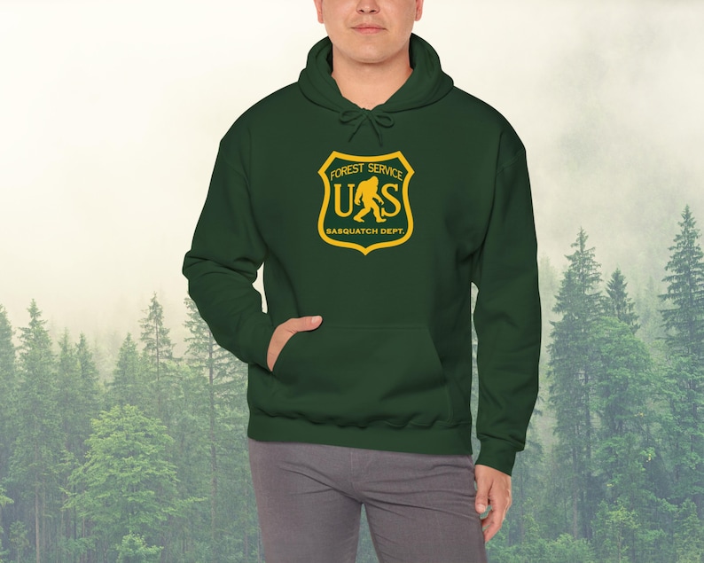 May include: A dark green hooded sweatshirt with a yellow and brown patch featuring a stylized image of a sasquatch and the text "Forest Service US Sasquatch Dept."