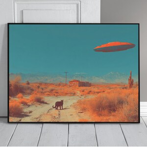 May include: Framed art print depicting a desert landscape with a black cat on a dirt road. An orange flying saucer hovers in the teal sky above a small building and mountains. The artwork has a retro, surreal vibe.