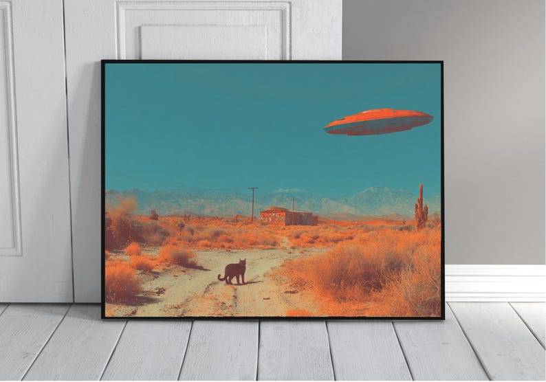 May include: Framed art print depicting a desert landscape with a black cat on a dirt road. An orange flying saucer hovers in the teal sky above a small building and mountains. The artwork has a retro, surreal vibe.