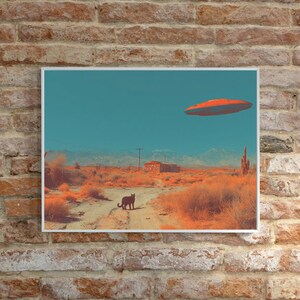 May include: A framed art print depicts a surreal desert scene. A black cat walks on a dirt road, with a red and orange UFO hovering in the turquoise sky. The landscape is rendered in warm orange and brown tones.