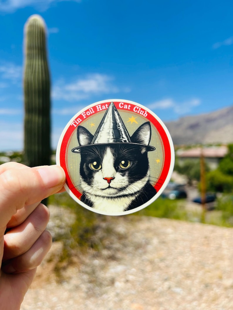 May include: A round sticker with a black and white cat wearing a silver tin foil hat. The sticker has a red border and the text "Tin Foil Hat Cat Club" in white.