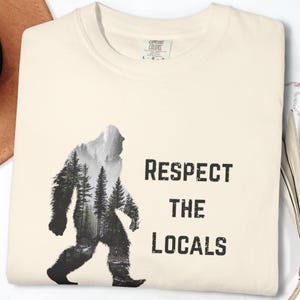 May include: Cream-colored sweatshirt featuring a black and white silhouette of Bigfoot filled with a forest scene. The text "RESPECT THE LOCALS" is printed in black, below the image. The sweatshirt has a crew neck and long sleeves.