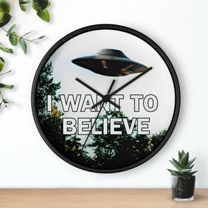 May include: A round black wall clock with a white face featuring a black UFO illustration above a forest scene. The text "I WANT TO BELIEVE" is printed in black. Black clock hands indicate the time.