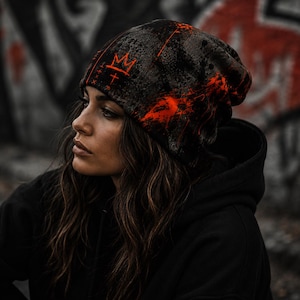 Close-up of woman wearing black and orange grunge beanie featuring distressed graffiti textures, orange crown artwork and paint splatter, alternative streetwear style.