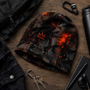 Black and orange grunge beanie flat lay with distressed graffiti crown design, paint splatter and charcoal textures styled with black streetwear accessories.