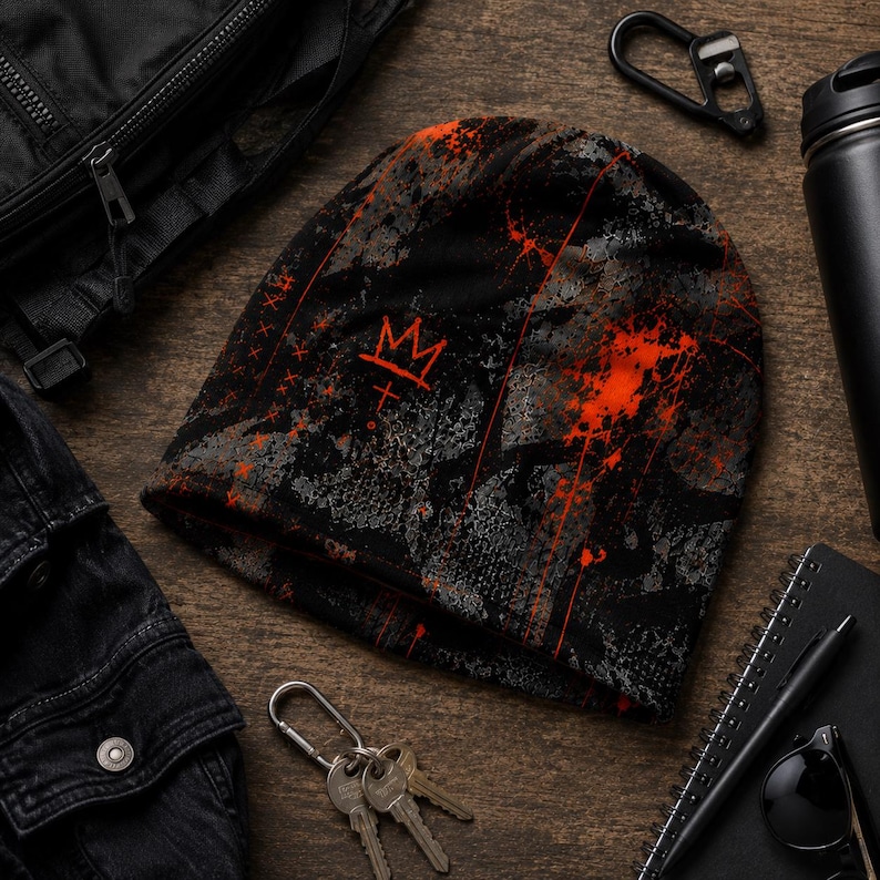 Black and orange grunge beanie flat lay with distressed graffiti crown design, paint splatter and charcoal textures styled with black streetwear accessories.