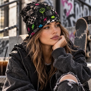 May include: A black beanie featuring a checkered pattern and neon-colored graphic prints including a smiley face, lightning bolts, and pixelated shapes. The fabric has a textured appearance with vibrant pink, green, and blue accents throughout the design.