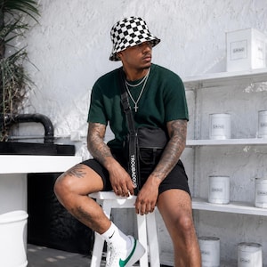 Peut inclure: Une personne portant un bob à carreaux noir et blanc, un t-shirt vert foncé à manches courtes et un short noir. Un sac bandoulière noir avec le mot "TROUBADOUR" est visible. La personne porte des chaussettes blanches et des baskets avec un logo vert.