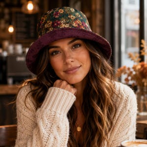 May include: A reversible bucket hat featuring a dark green crown with a vibrant multicolored floral pattern and a solid deep purple brim. The hat is constructed with a structured design and a wide, downward-sloping brim that frames the face.