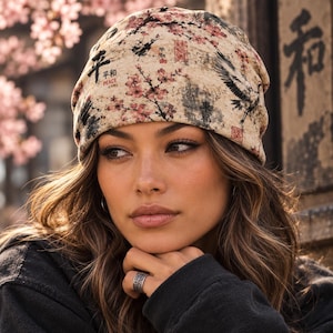 May include: A beige beanie featuring a printed pattern of pink cherry blossoms, black flying cranes, and Japanese calligraphy characters including the word PEACE. The fabric has a textured appearance with various red stamp-like markings.