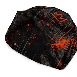 Folded black and orange grunge beanie displaying distressed crown artwork, graffiti symbols, paint splatter and dark urban streetwear pattern.