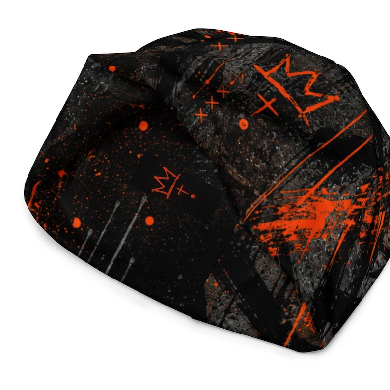 Folded black and orange grunge beanie displaying distressed crown artwork, graffiti symbols, paint splatter and dark urban streetwear pattern.