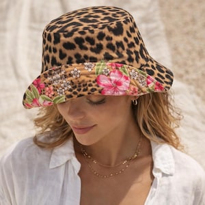 Könnte beinhalten: Ein Bucket Hat mit Leopardenmuster und floral gemustertem Rand. Der Hut hat ein braunes und schwarzes Fleckenmuster auf der Krone und ein farbenfrohes Blumendesign mit rosa und weißen Blumen auf dem Rand. Ein stilvolles Accessoire.