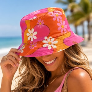 May include: A bucket hat featuring a vibrant pattern of white daisies against a background of wavy orange and pink sections. The hat has a downward-sloping brim and a rounded crown, constructed from fabric with a colorful floral print design.