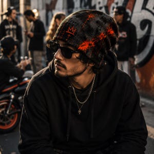 Man wearing black and orange grunge beanie with distressed graffiti crown design, styled with black hoodie, sunglasses and chains in an urban street art setting, alternative men’s streetwear look.