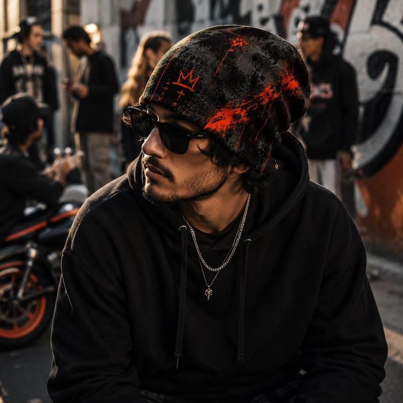 Man wearing black and orange grunge beanie with distressed graffiti crown design, styled with black hoodie, sunglasses and chains in an urban street art setting, alternative men’s streetwear look.