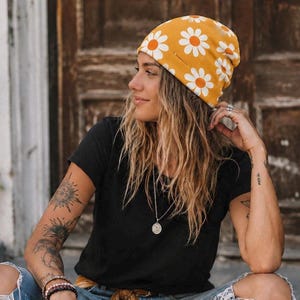 May include: A mustard yellow beanie featuring a repeating white daisy pattern with orange centers. The hat is made of a soft fabric and is worn in a slouchy style, covering the top of the head with a rounded silhouette and a fitted band.
