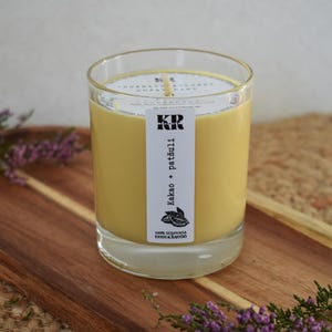Può includere: Una candela di cera di soia gialla in un contenitore di vetro trasparente. La candela ha un'etichetta bianca con il testo "kaka + patšuli" e un disegno di chicchi di cacao. La candela è su un vassoio di legno con fiori viola.